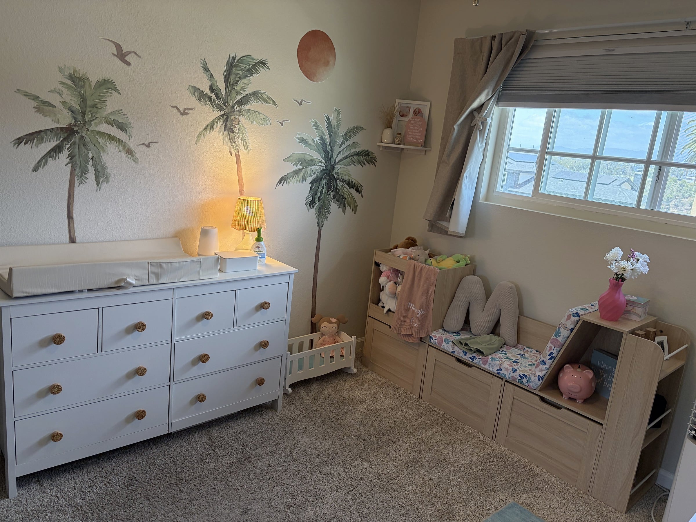 A child's bedroom with a white seven-drawer dresser, palm tree wall decals, a toy storage unit, and a window with beige curtains