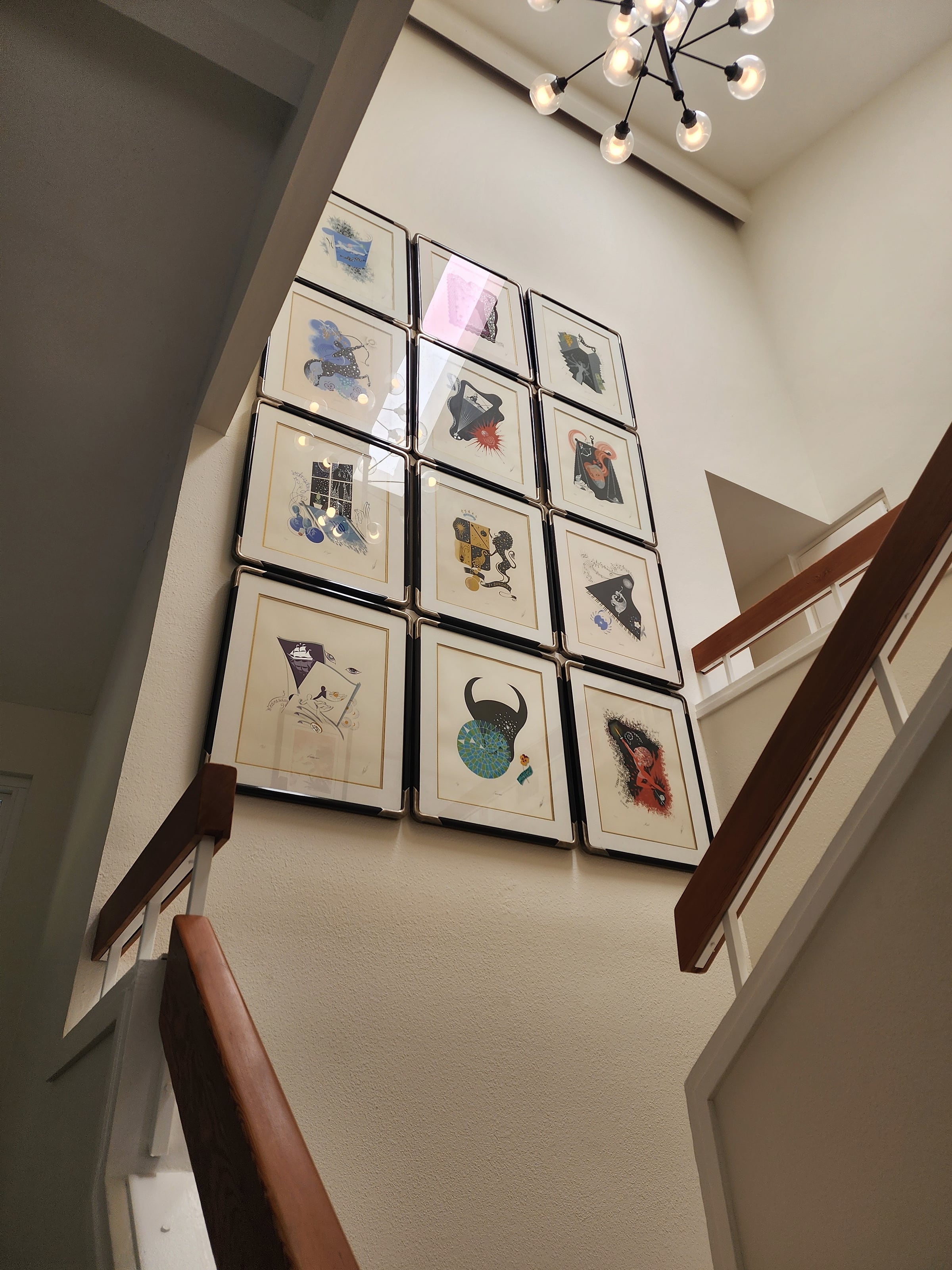 Twelve black-framed colorful prints arranged in a 3×4 grid on a tall white stairwell wall, with wood railings and a sputnik chandelier above