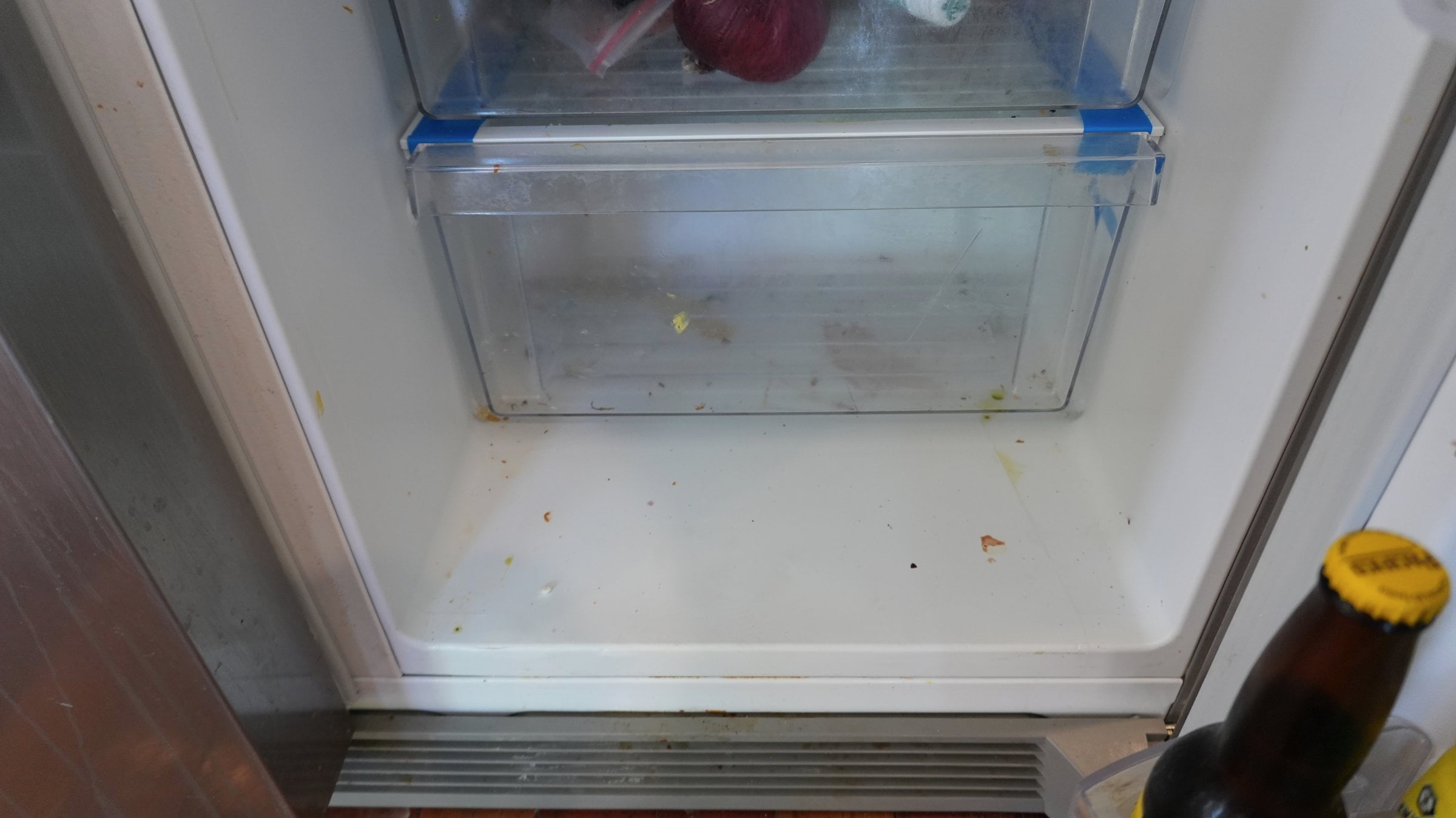 Open refrigerator bottom shelf showing a dirty crisper drawer and floor with food debris and stains