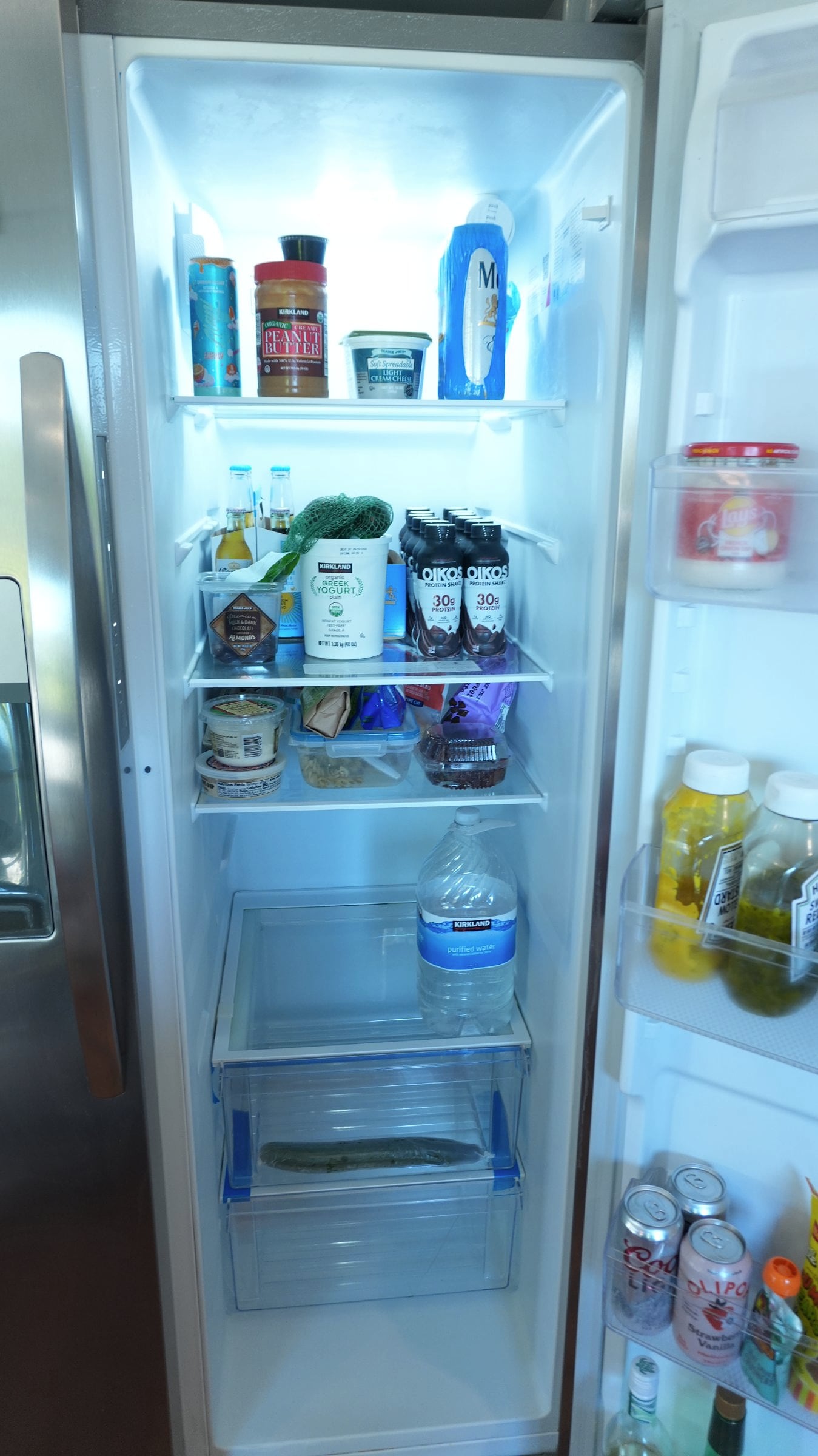 Open stainless-steel refrigerator holding peanut butter, Greek yogurt, Oikos protein shakes, a water jug, and condiments on door shelves