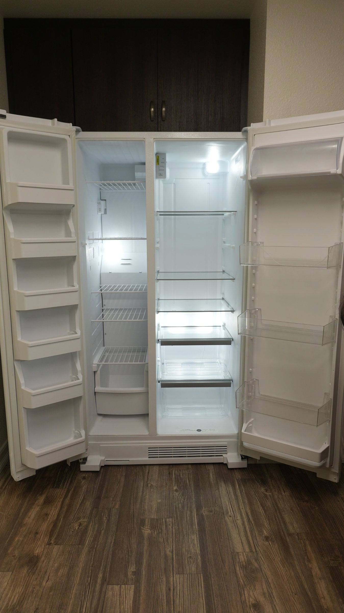 White side-by-side refrigerator with both doors open, showing empty lit shelves and door bins, beside dark wood cabinets