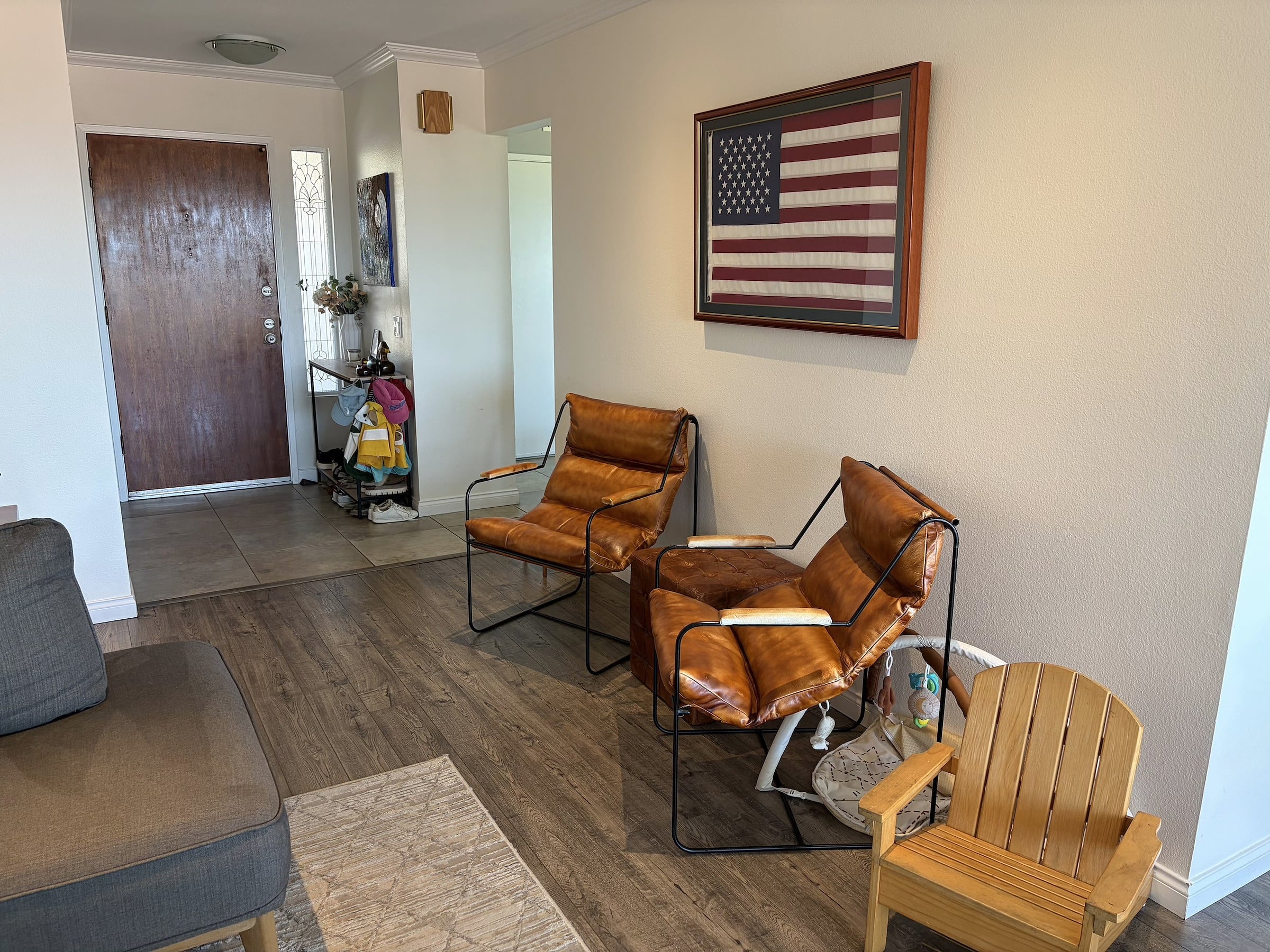 Two tan leather sling chairs and a small wooden chair sit on wood floors near a framed American flag, with a front door visible in the ba…