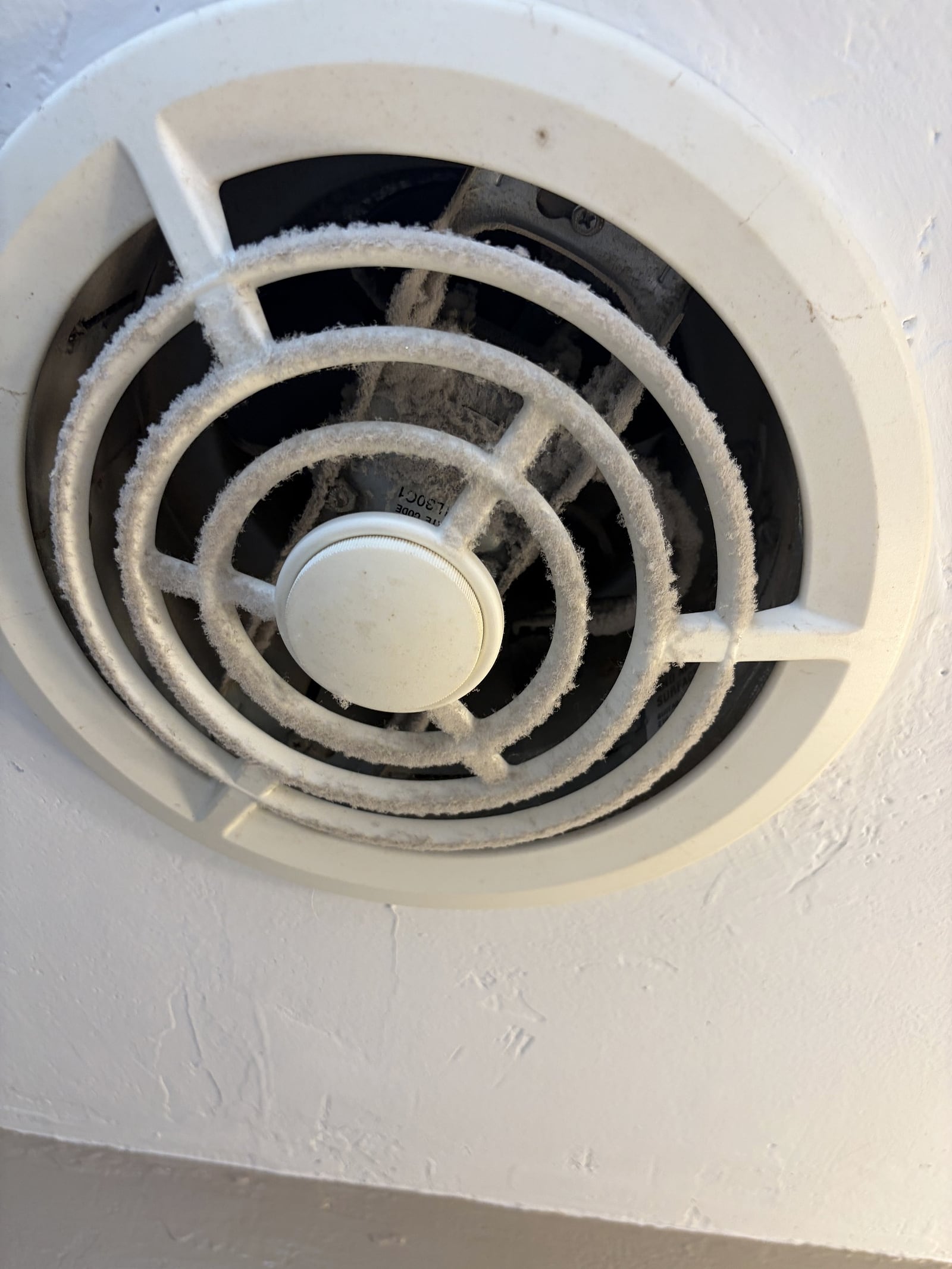 A round white bathroom exhaust fan vent on a ceiling, heavily coated in gray dust and lint buildup