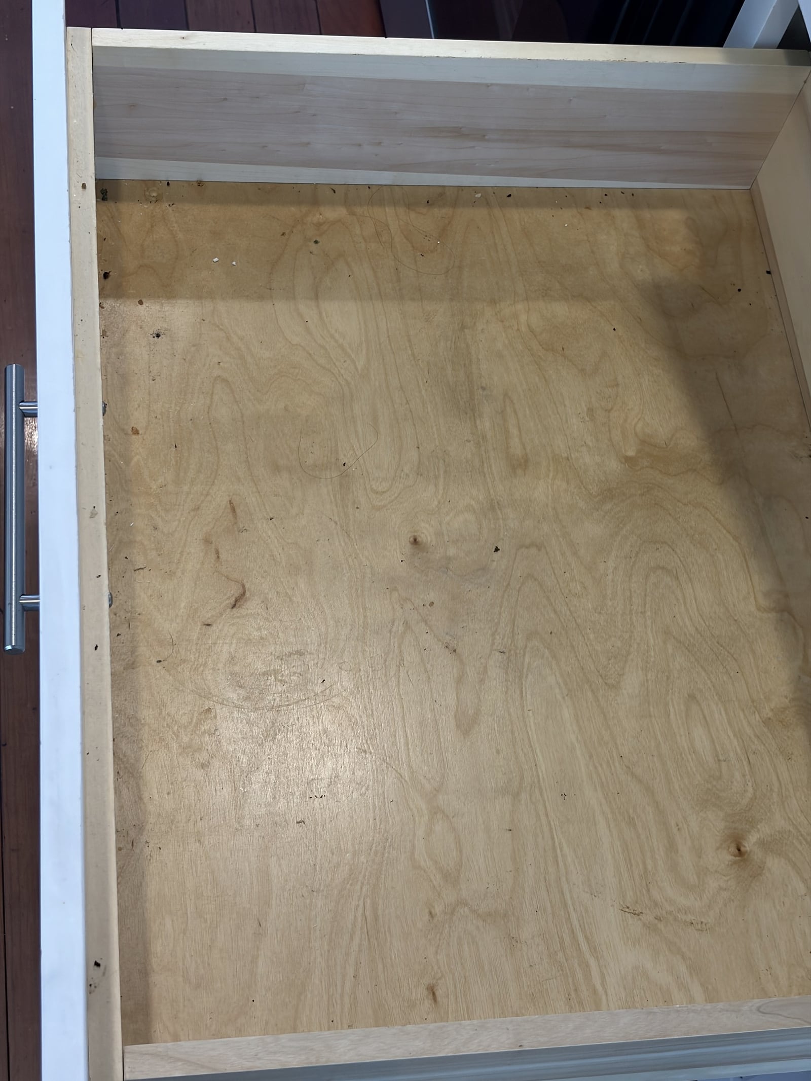 Open empty wooden drawer with a silver handle showing dirt and debris scattered across the plywood base