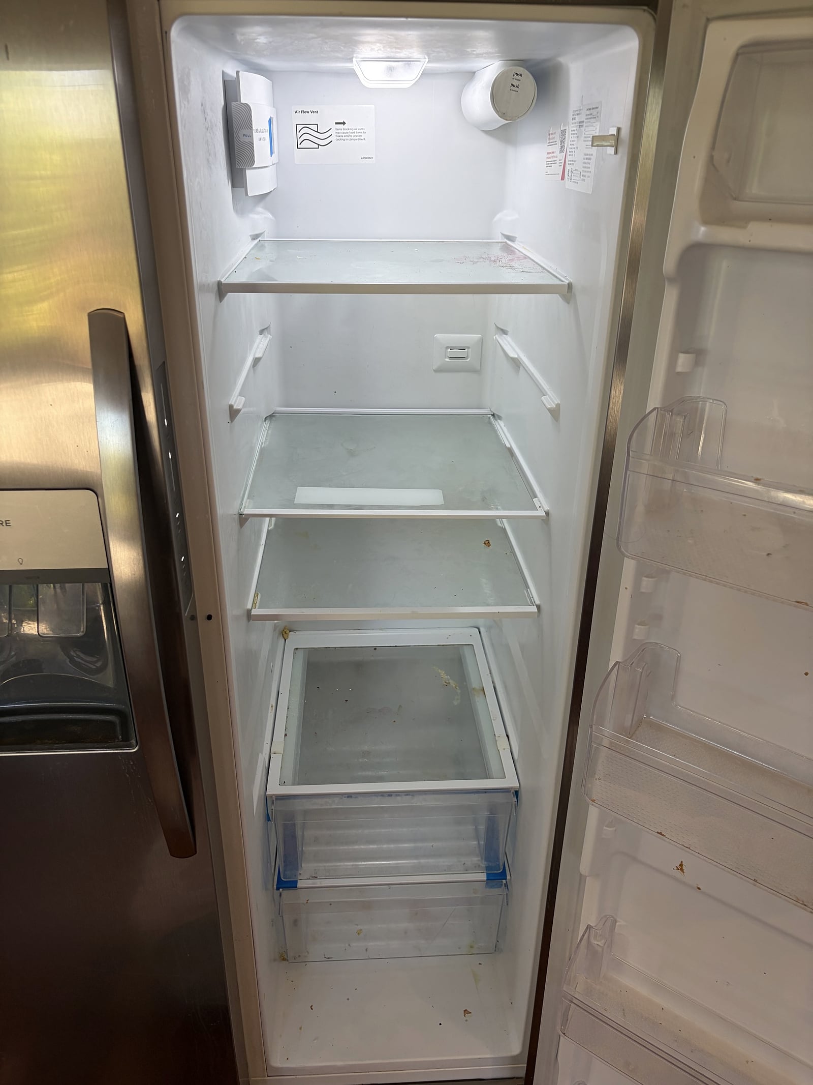Open stainless steel refrigerator interior with empty glass shelves, two clear crisper drawers, and visible food spills on the floor and…