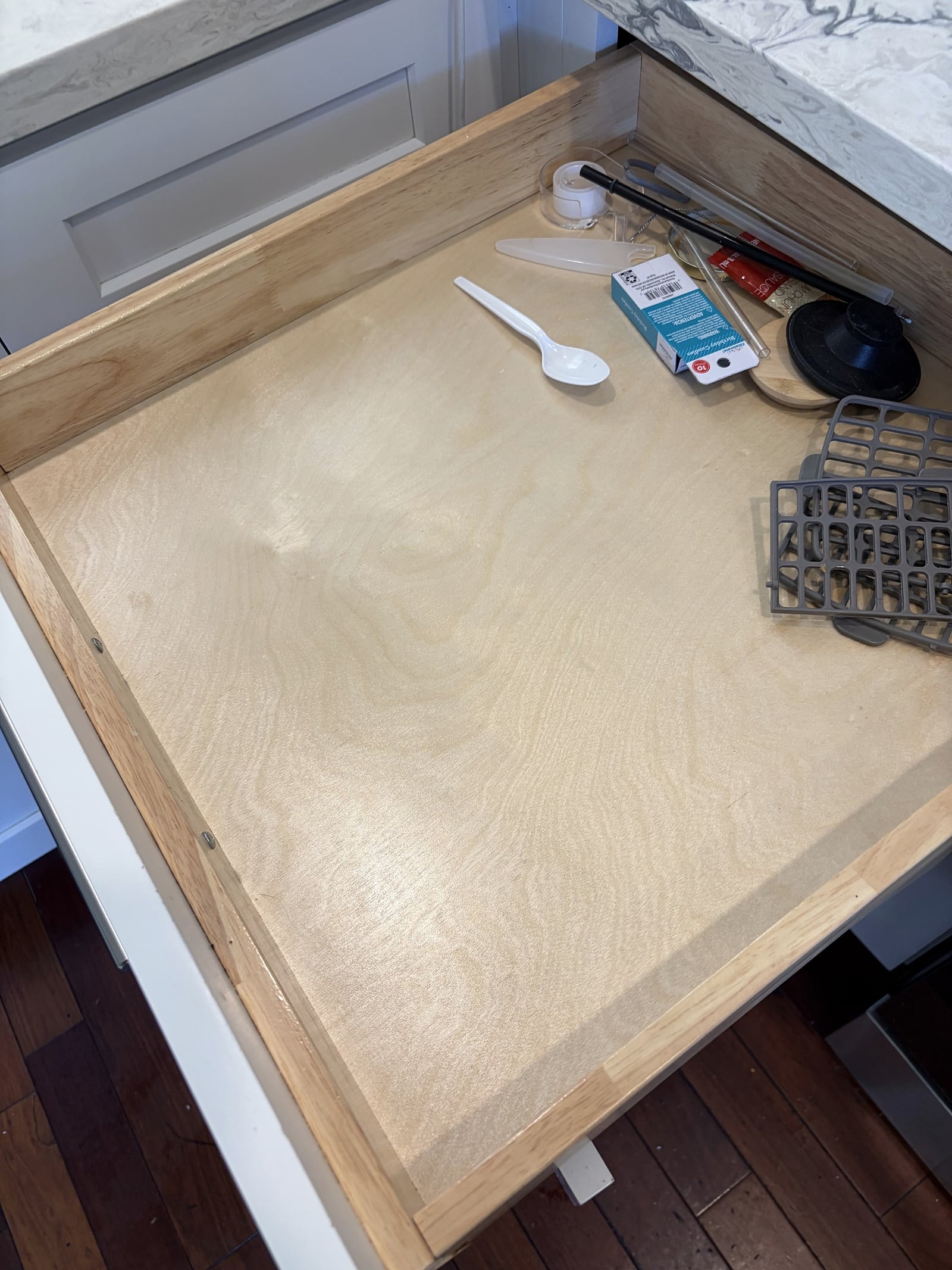 Open kitchen drawer with a bare wood base holding a plastic spoon, small box, tongs, and a gray trivet in one corner