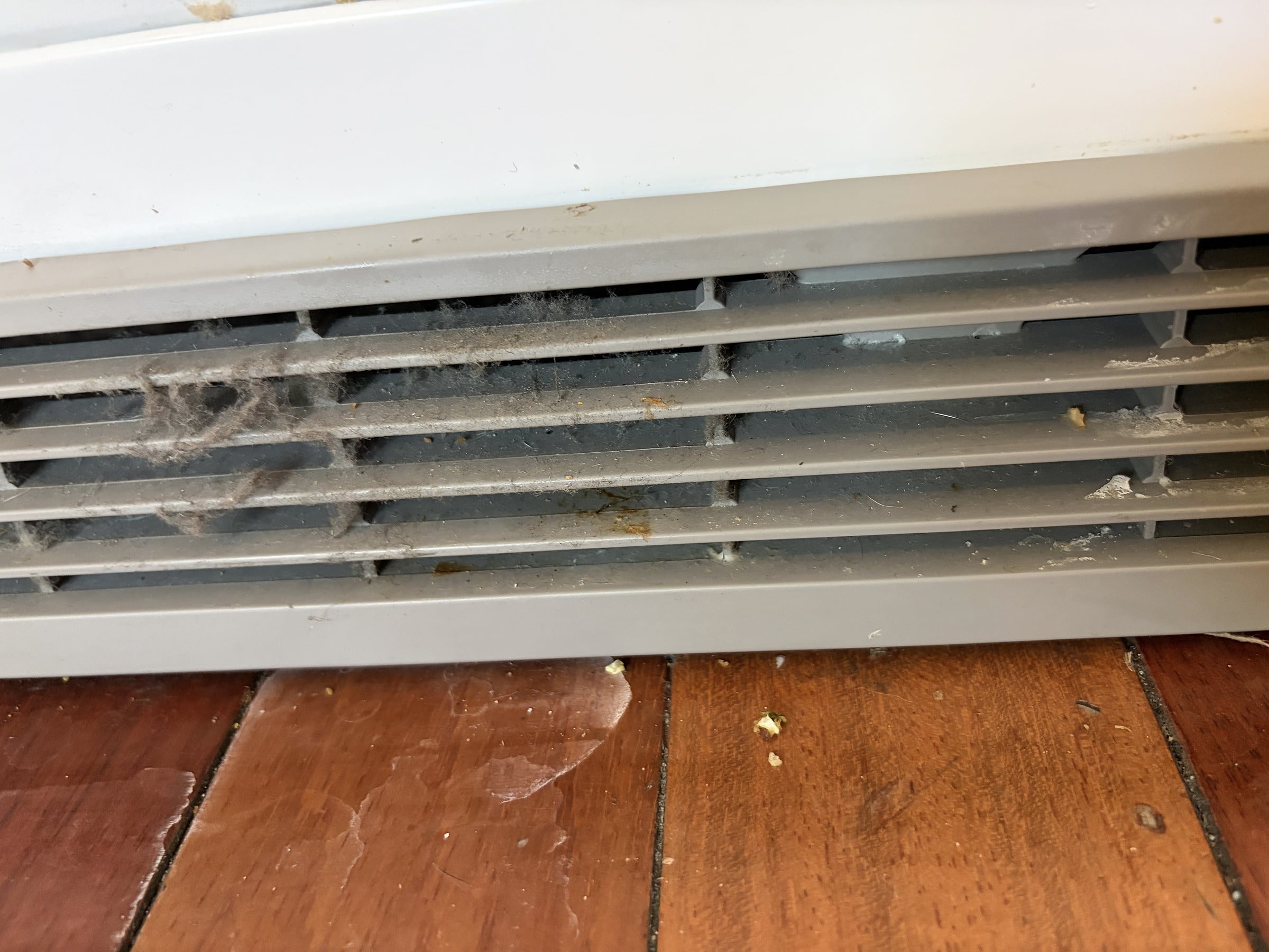 A white floor-level air vent caked with dust and debris, sitting above a scratched hardwood floor with scattered dirt particles