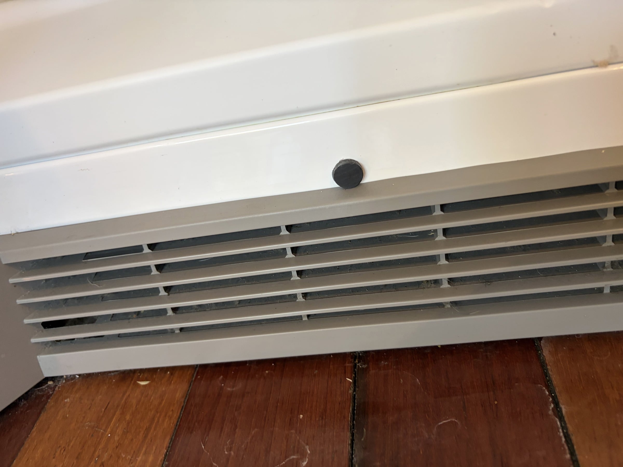 White baseboard heating vent cover with visible dust and debris on wood floor beneath it