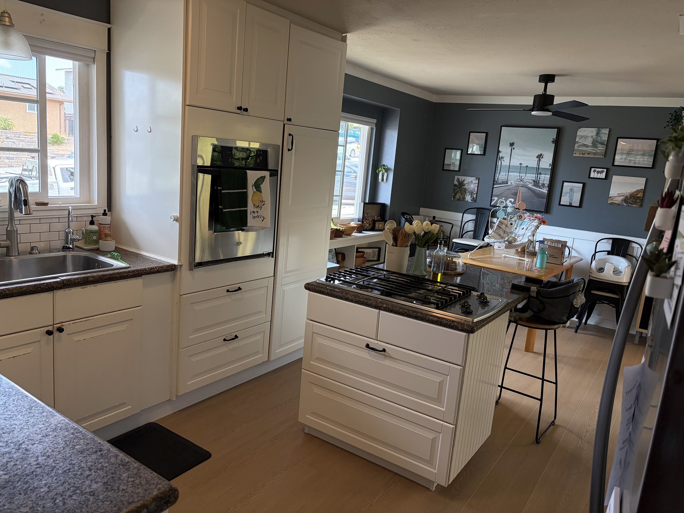 White kitchen with a center island, gas cooktop, stainless wall oven, and sink, opening into a gray dining area with framed wall art