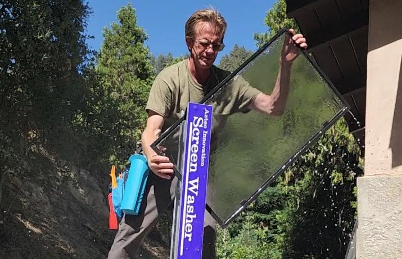 Man holding a wet window screen with an Aztec Innovation Screen Washer tool attached, outdoors with trees in the background