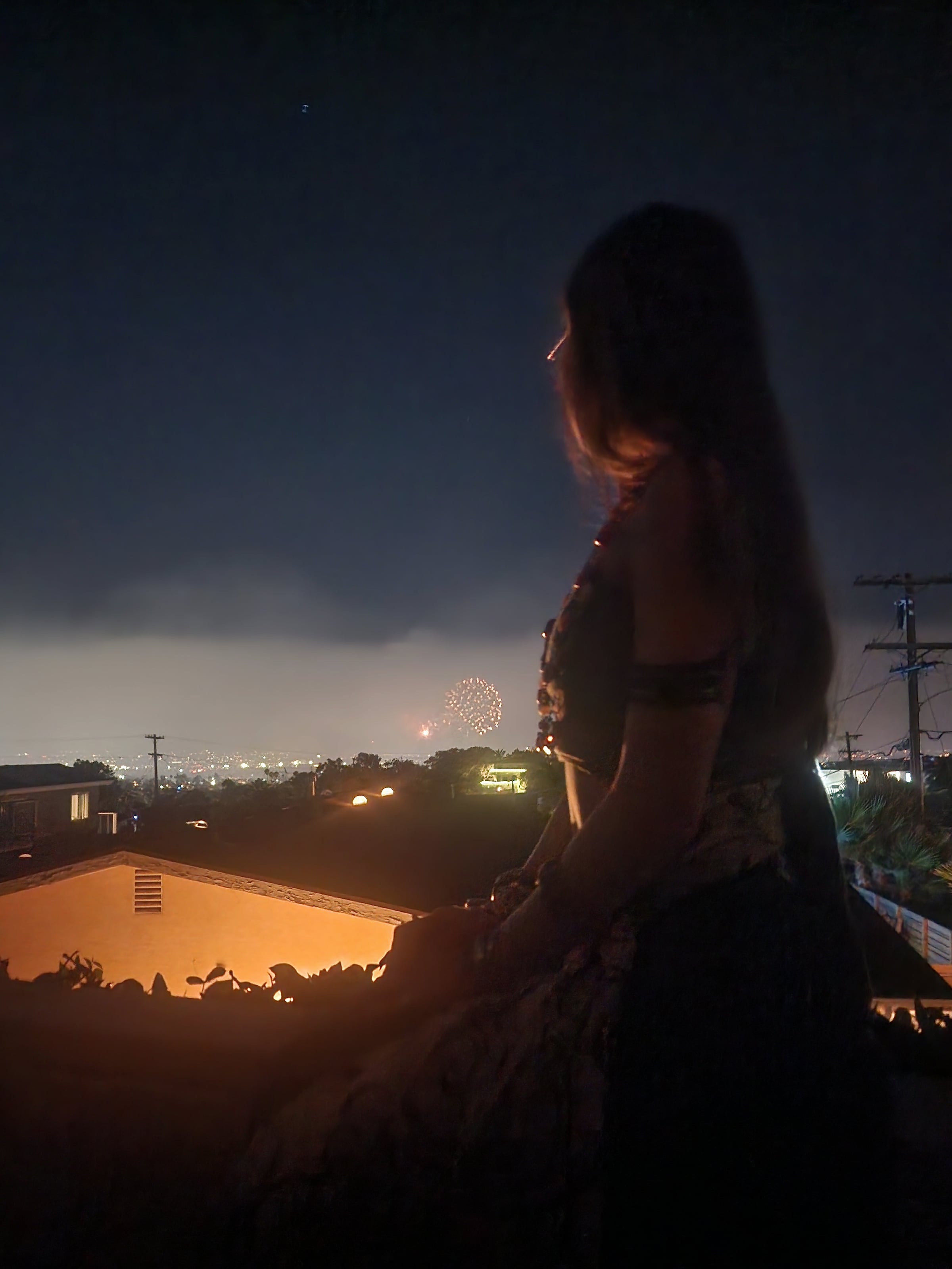 A woman with long hair stands on a rooftop at night watching fireworks burst over a residential neighborhood